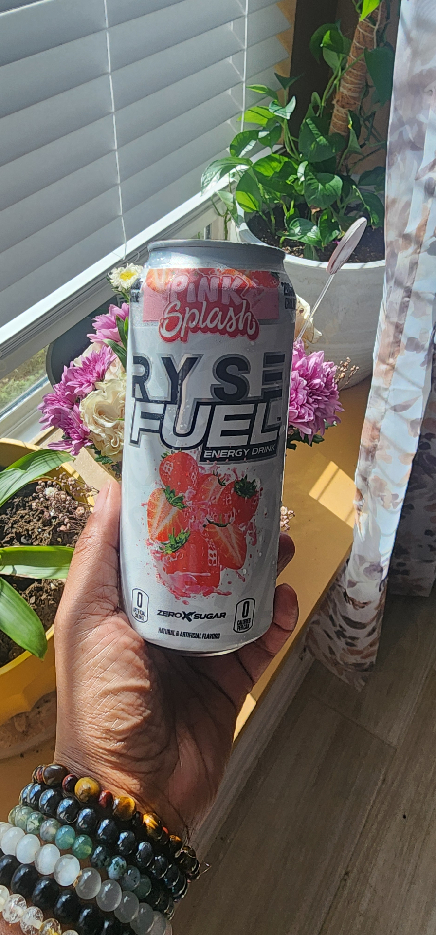 Hand holding a RYSE Fuel Pink Splash energy drink can in front of a sunny window with plants and flowers